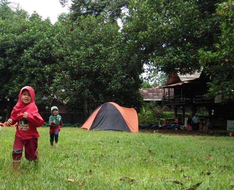Dua siswa berlari di halaman berumput dengan tenda kemah di belakangnya